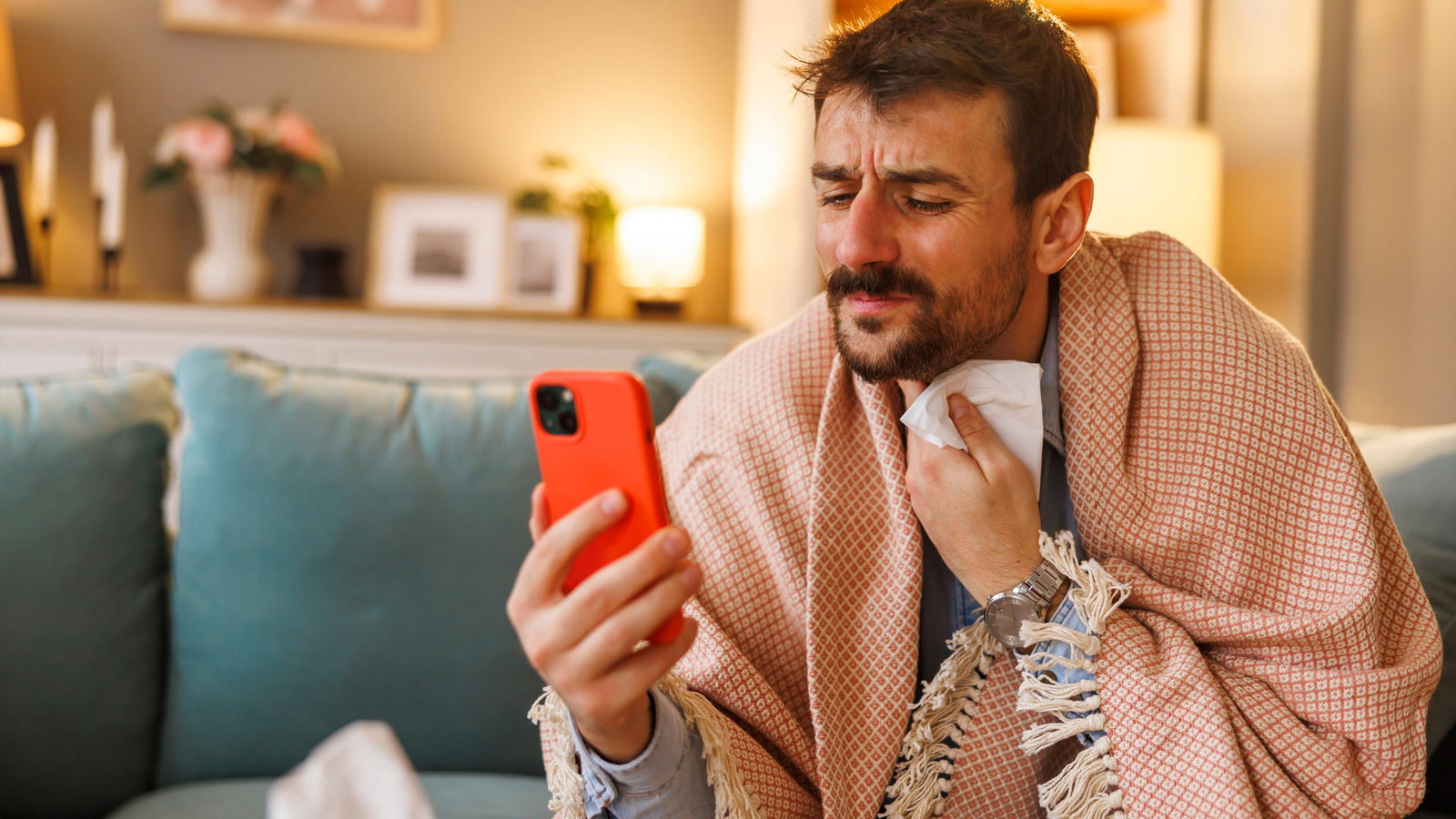 Kranker Mann sitzt auf der Couch mit Decke und schaut in sein Smartphone